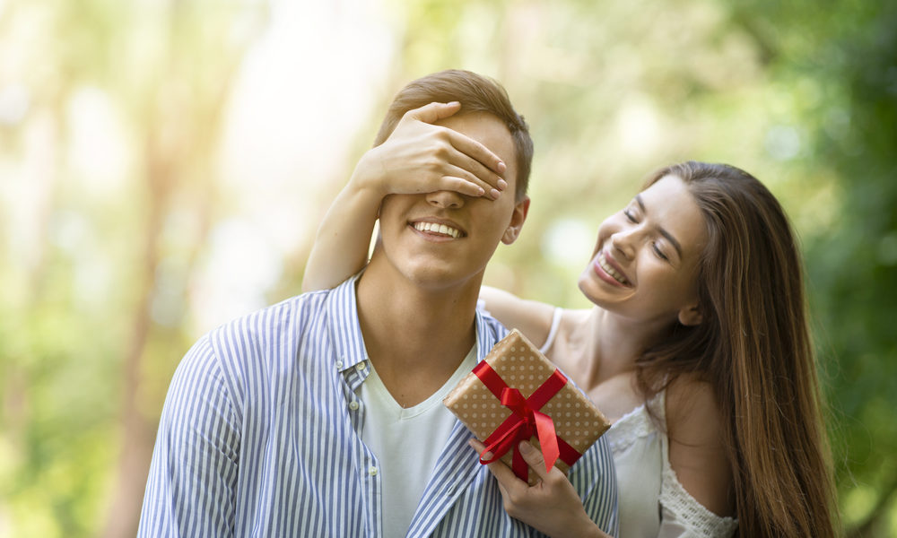 Attractive young girl closing her boyfriend's eyes and surprising him with birthday gift at park