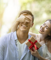 Attractive young girl closing her boyfriend's eyes and surprising him with birthday gift at park