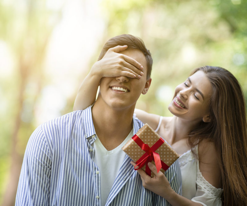 Attractive young girl closing her boyfriend's eyes and surprising him with birthday gift at park